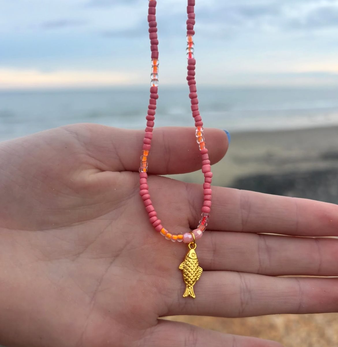 Pink beaded fish necklace held in an open hand against an ocean background